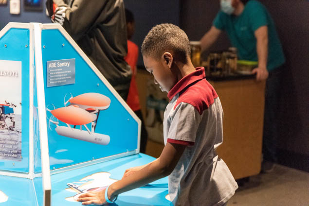 young boy in grey shirt stands next to interactive with submarine on it