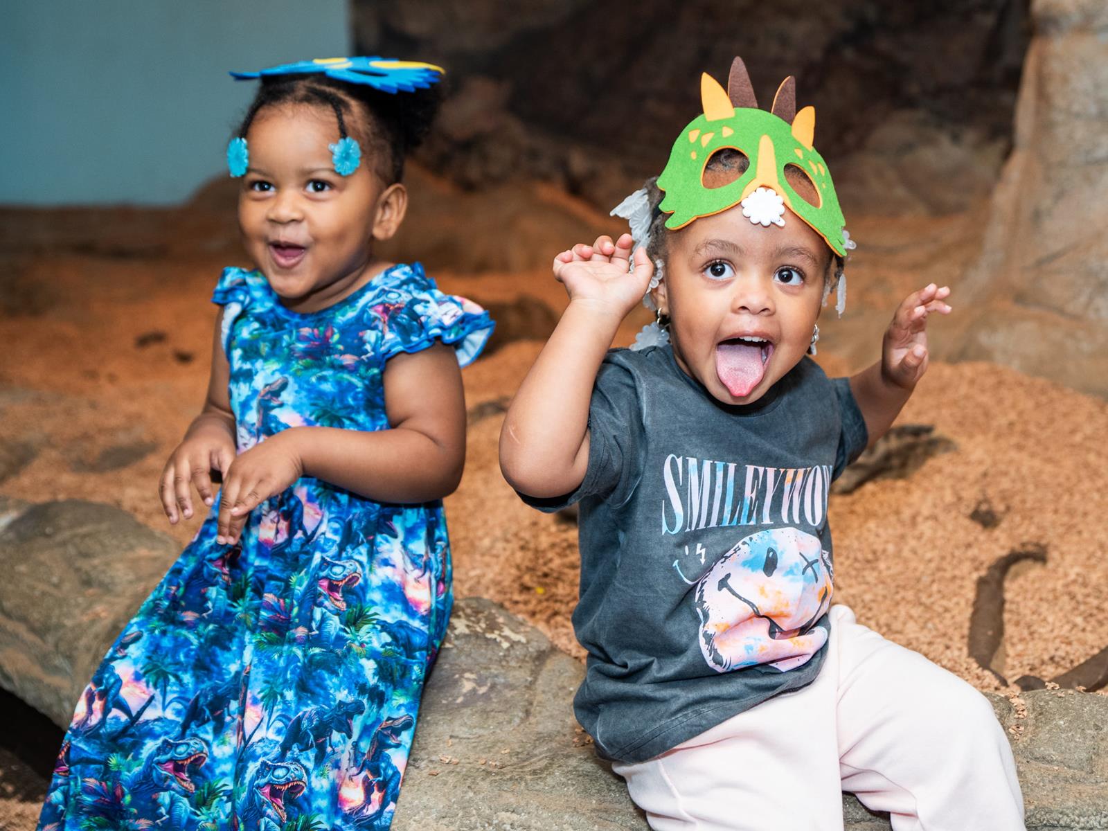 Two children posing for a photo in the big dig, both wearing dinosaur masks on their head.
