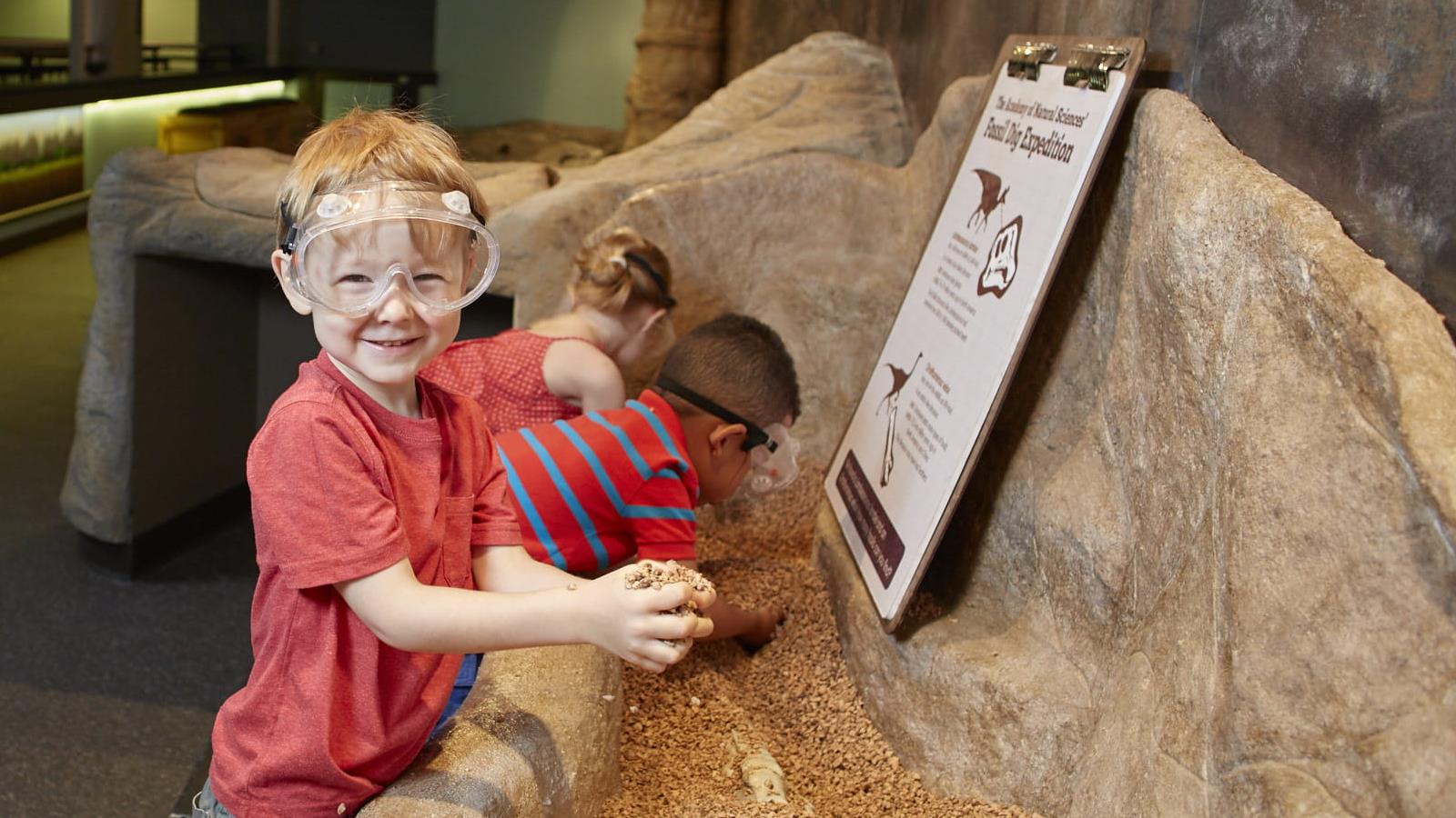 Three children looking for fossils in the big dig, all wearing goggles.