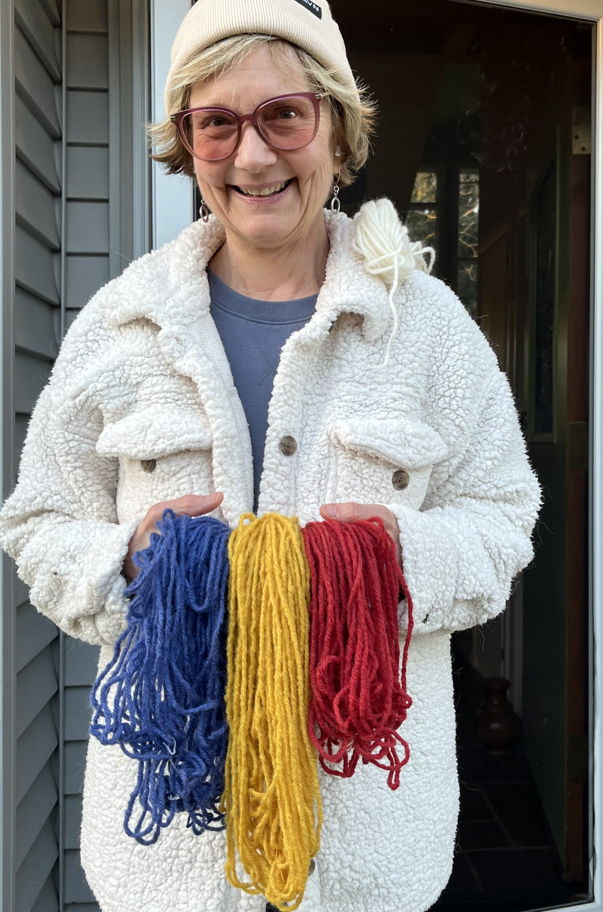 A woman wearing a white coat and hat, holding three fabrics of different colors, red, blue, and yellow.