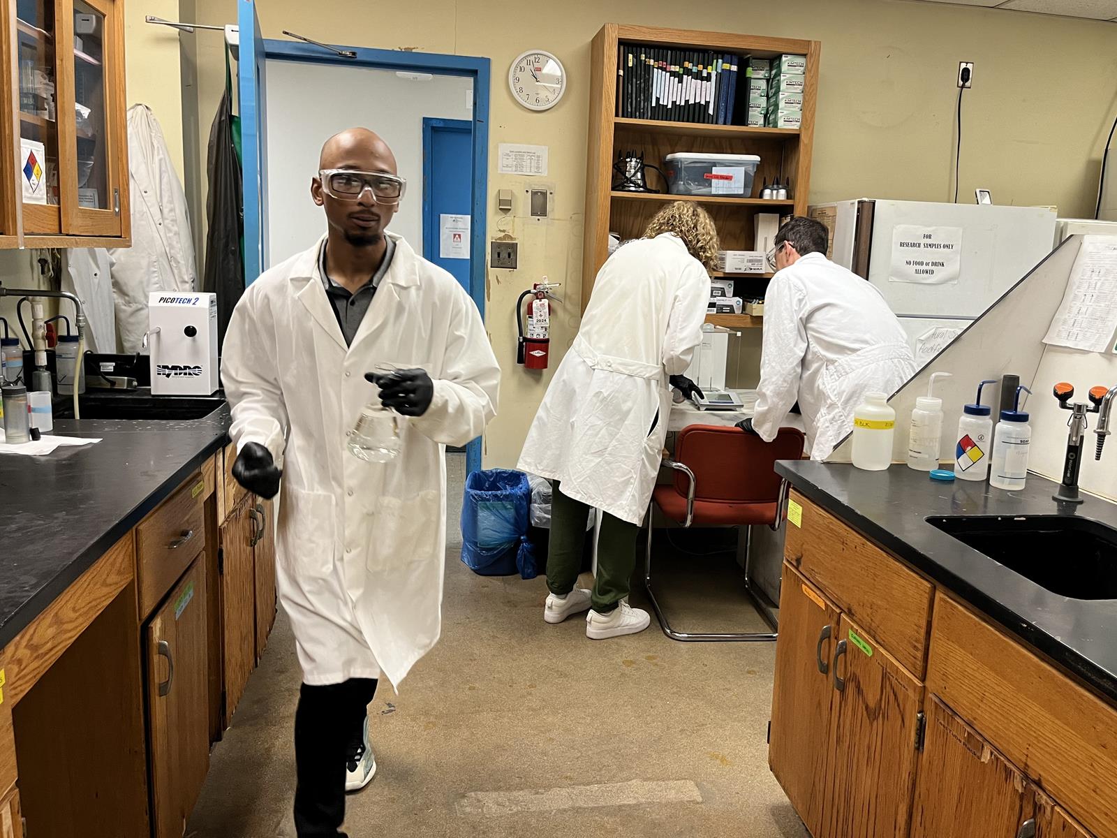 Three volunteers working in a lab, two weighing a specimen on a scale and the other carrying a beaker filled with a clear liquid.