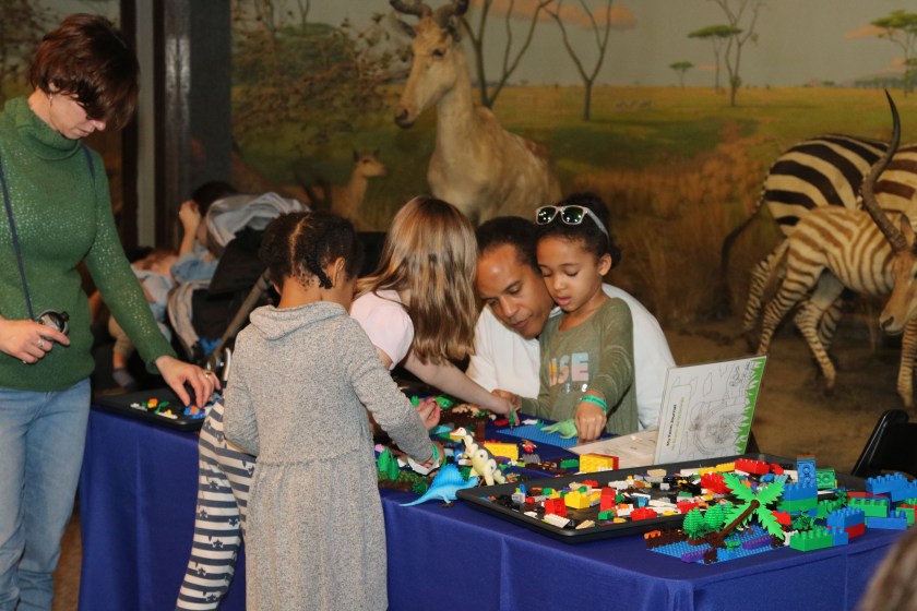 Two families play with legos and plastic dinosaurs at long table