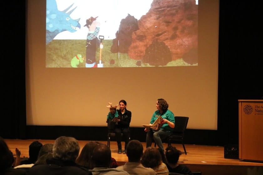 Two female educators onstage reading a story with moose puppet; story projected onscreen.