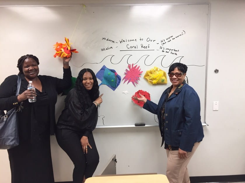Teachers holding colorful coral reef art in front of whiteboard