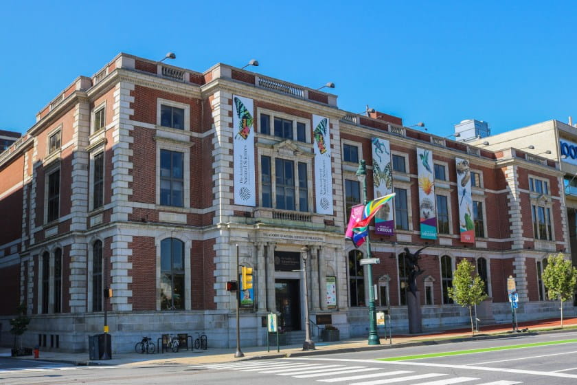 Image of the Academy Building on a summer day, three stories tall, red brick with white stone columns and window surrounds. Banners displaying butterflies and dinosaurs decorate the front of the building