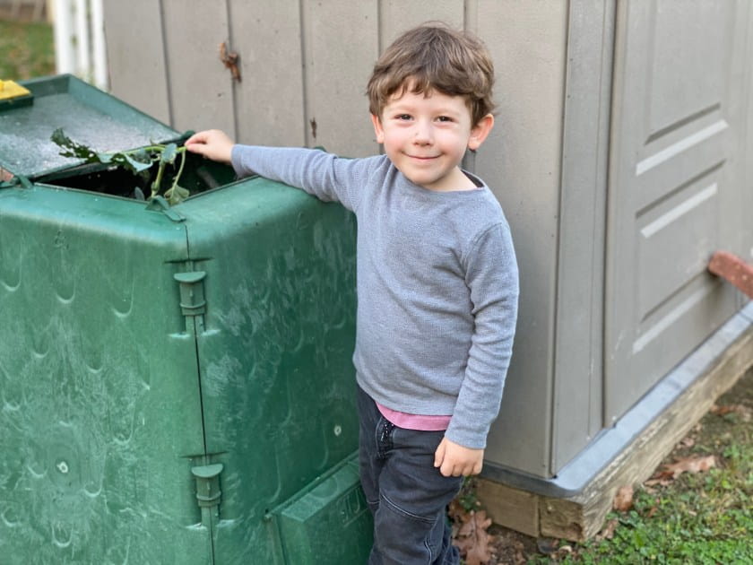 Three year old boy with brown hair, gray shirt and black pants places bird-eaten broccoli start into bright green compost bin that is almost as tall as he is