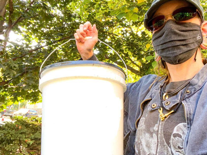 30-something adult wearing hat, sunglasses, face mask and denim jacket with gray t-shirt holds up white compost bucket against background of green treetops