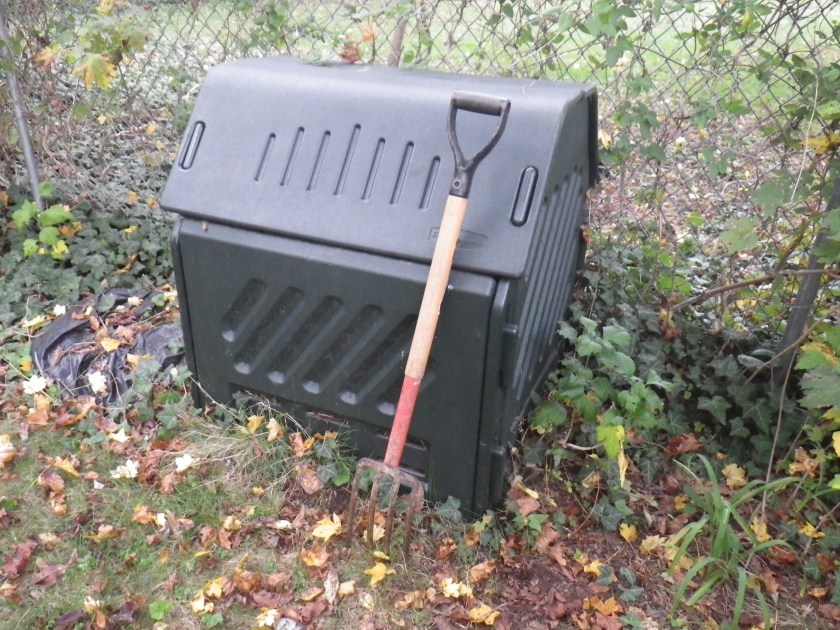 Black compost bin about three feet high with wooden handled small pitchfork leaning against it, up against a chain link fence with greens growing on it