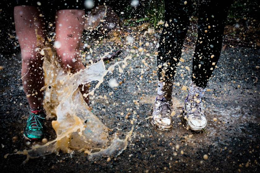 Two people splashing in muddy puddle. One wears shorts and teal sneakers, the other wears black leggings, white socks and white sneakers