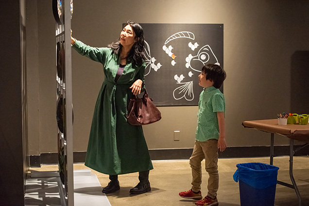 A mother in a dark green dress showing her son who is wearing a gingham shirt, an activitiy in the SKIN exhibit.