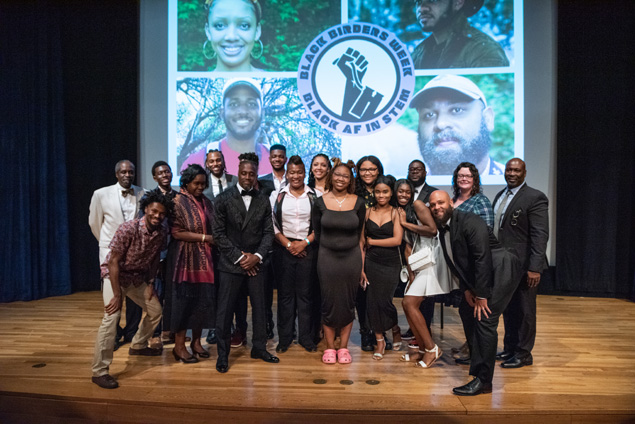 The Black Birders and others stand on the stage of the auditorium posing for the camera.