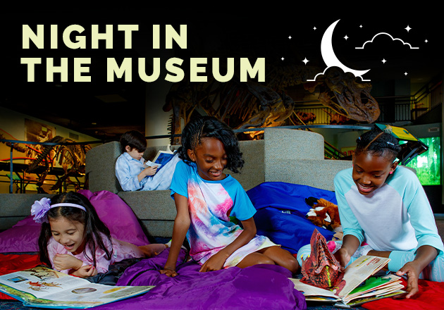 Four children wearing pajamas are reading books in dinosaur hall.