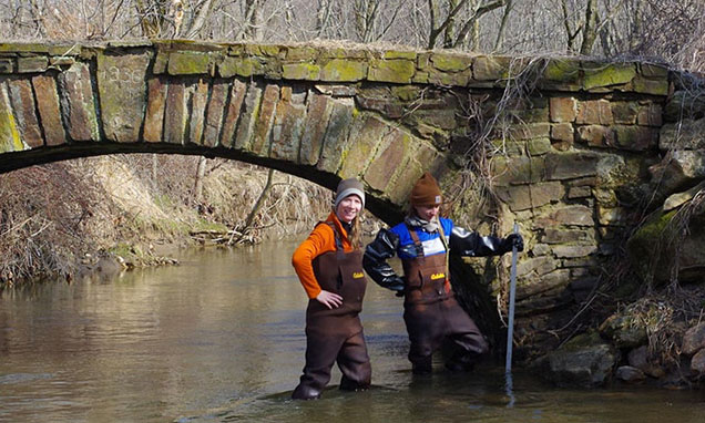 two scientists in gaiters standing in stream under old cement bridge