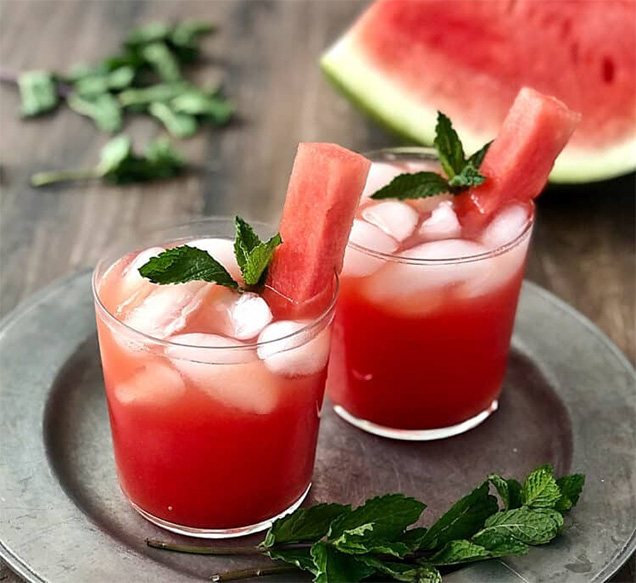 red drink in glass with ice and green garnish next to slice of watermelon