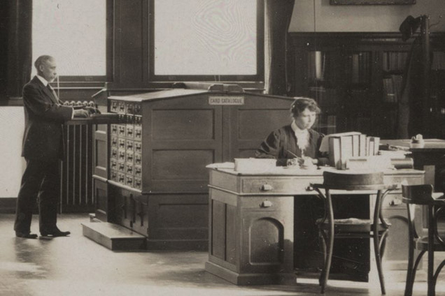 Sepia tone photo of woman sitting at desk with man at card catalog