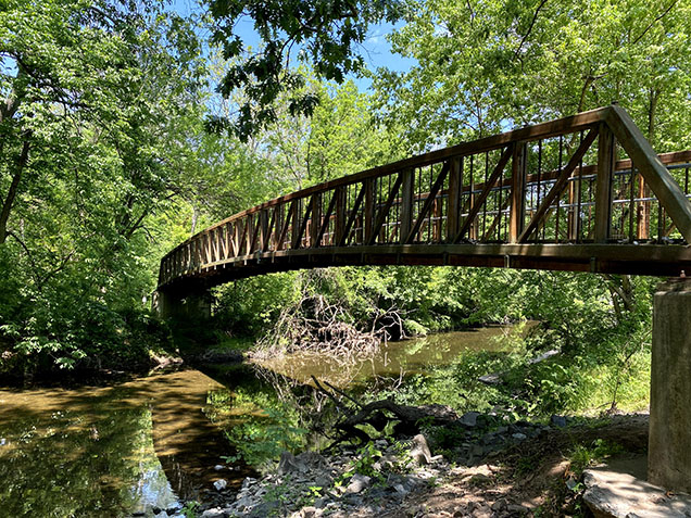 metal bridge over creek with green trees and blue sky