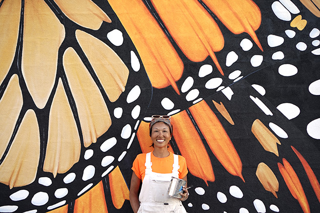woman in orange shirt and white overalls stands in front of painting of orange and black butterfly wings